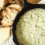 Spinach and artichoke dip in a cast iron skillet with toasted bread to the left and a chunk of parmesan.