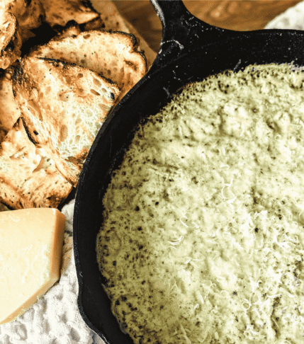 Spinach and artichoke dip in a cast iron skillet with toasted bread to the left and a chunk of parmesan.