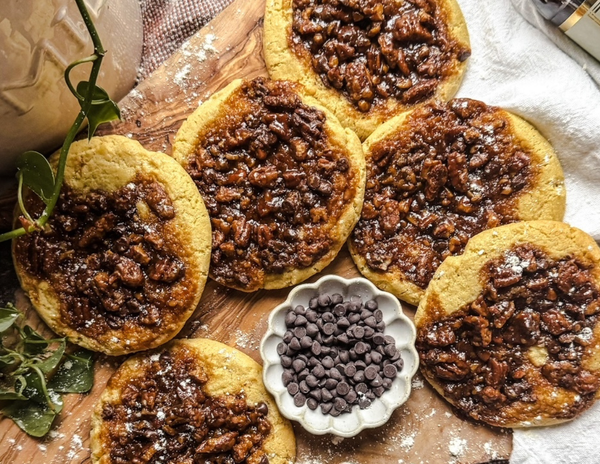 Pecan Pie Cookies sitting on a cutting board near a small container of chocolate chips in the sun