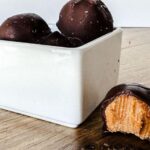 Crunchy peanut butter bombs coated in chocolate, sitting in a white bowl with a wooden background.