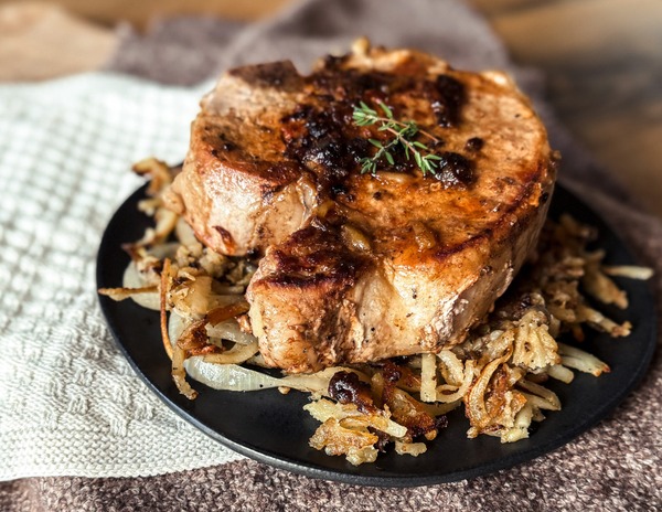 A neautral back drop with a black circular plated covering most of the photo. A large seared pork chop is sitting on top of hash browns on the plate.
