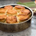 A skillet with a chicken gravy below and biscuits on top. Celery and carrots in the backdrop.