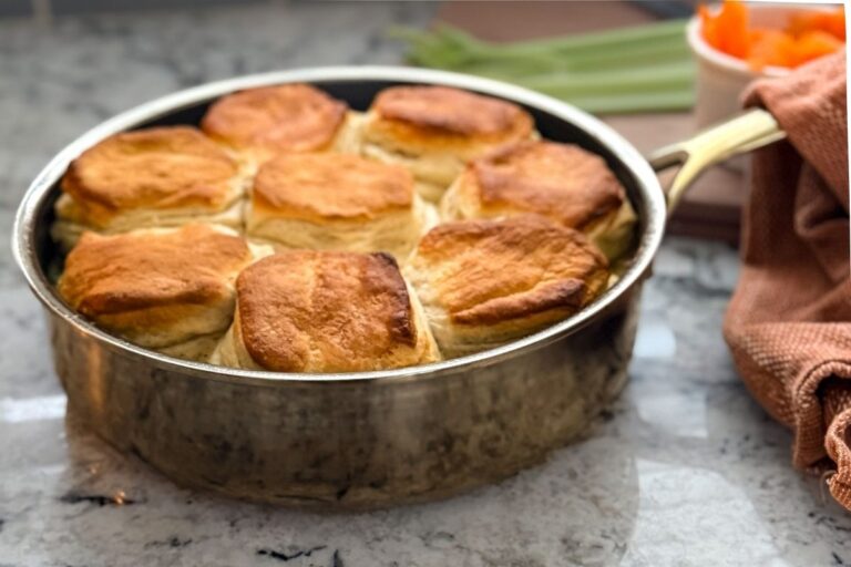 A skillet with a chicken gravy below and biscuits on top. Celery and carrots in the backdrop.