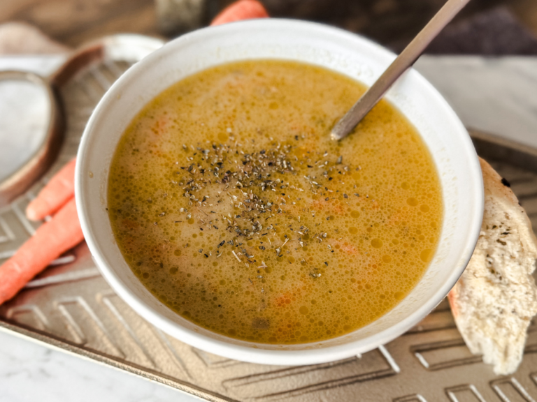 Gold tray with a white bowl on top. Orange carrots on the left side and the bowl is filled with an orange and yellow cheddar broccoli soup. A gold spoon is placed in the soup. Spices are sprinkled on top of the soup.