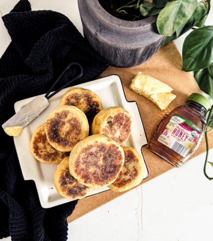 Pile of fresh baked english muffins sitting on a white plate near a plate tea towel, some butter, and a container of honey