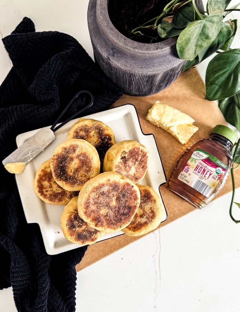 Pile of fresh baked english muffins sitting on a white plate near a plate tea towel, some butter, and a container of honey