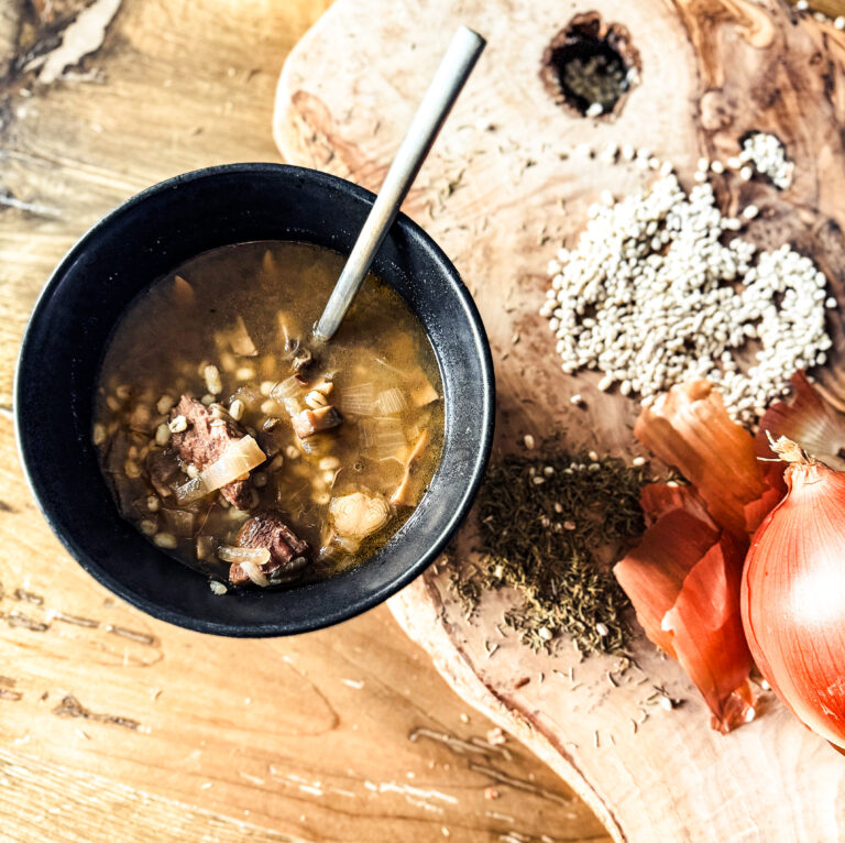 Beef and Barley Soup in a black bowl with gold spoon. A wooden background has barley sprinkled and onion onion.