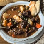 Beef Stew in a scalloped bowl with a scalloped plate underneath. There is a baguette in the background and a torn piece of the baguette dipped in the stew.