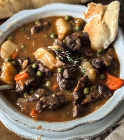 Beef Stew in a scalloped bowl with a scalloped plate underneath. There is a baguette in the background and a torn piece of the baguette dipped in the stew.