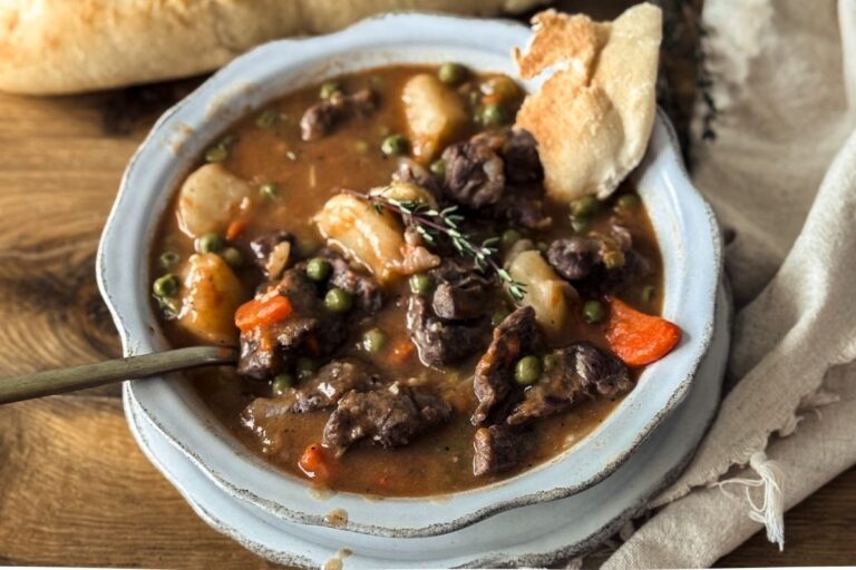 Beef Stew in a scalloped bowl with a scalloped plate underneath. There is a baguette in the background and a torn piece of the baguette dipped in the stew.