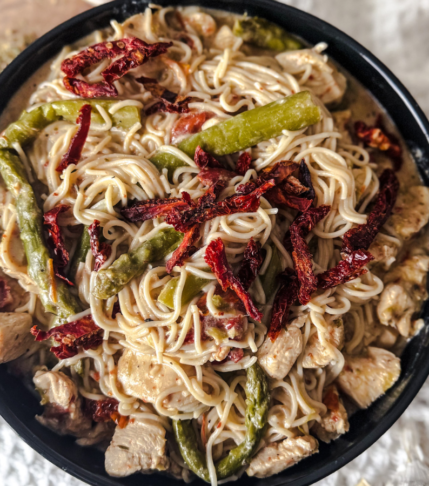 Black rimmed dish with wooden background and white cloth behind. The dish is filled with ramen and green asparagus, red sundried tomatoes and ramen topped with sesame seeds.