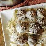 Slow Cooker Beef and Noodles on a white square plate. Gold tongs life a meatball. The backdrop is wooden with a pot to the top left.