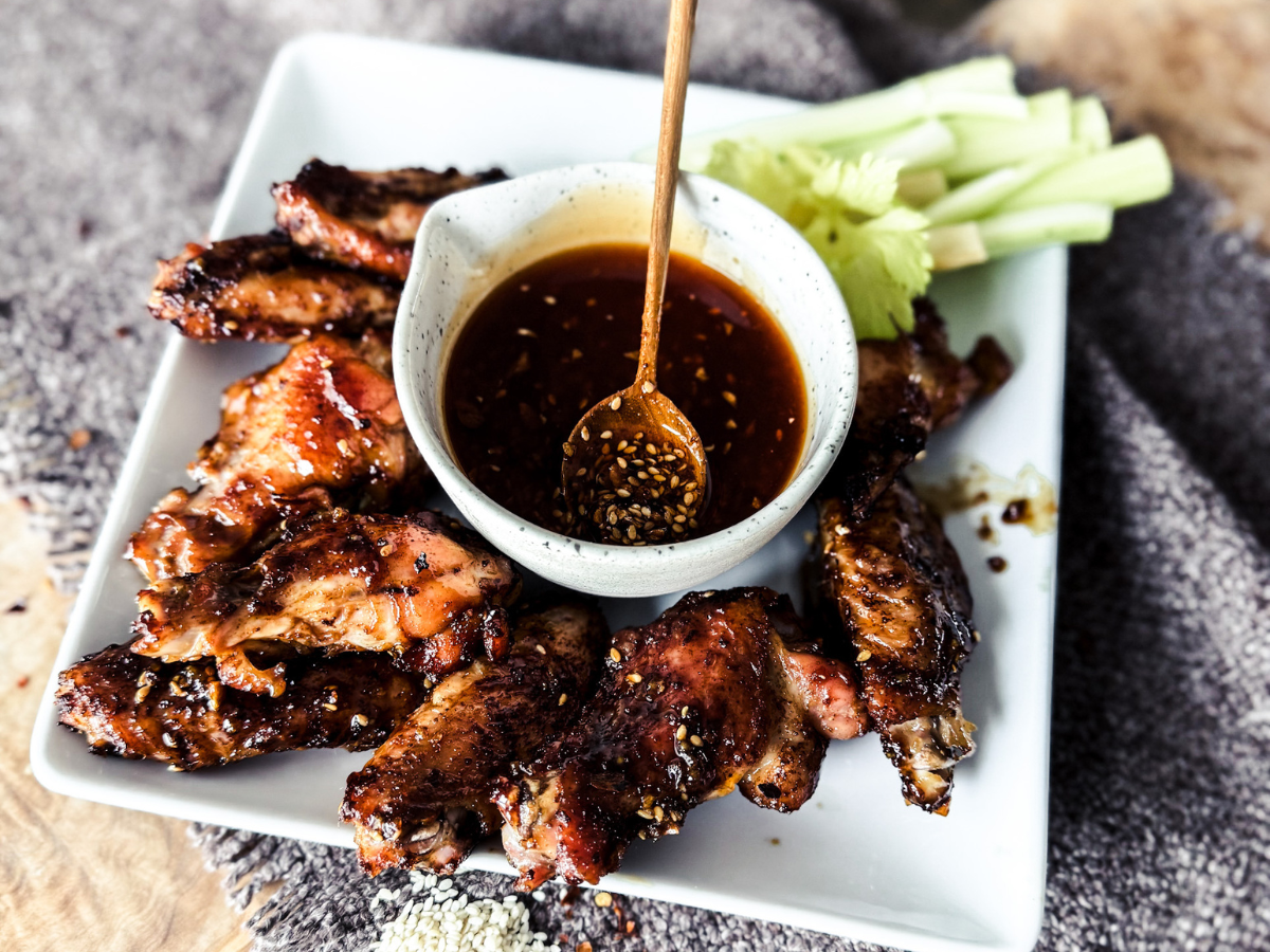 Medium brown textured and wood background. White square plate with chili sauce in the middle in a small white bowl. The bowl is surround by chicken wings with a little nit of celery.