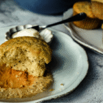 A half eaten poppyseed muffin on a shite scalloped dish with a small scalloped tray of butter sitting behind. 3 muffins piled to the right