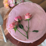 Circular vanilla cake covered in pink icing with 3 roses placedon a wooden cake stand.