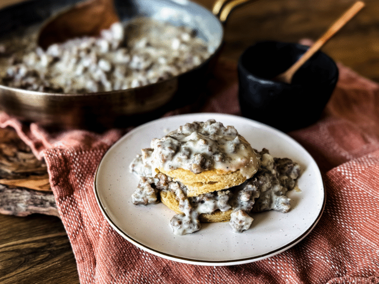 Biscuit topped with gravy on a white plate. The background is orange and wooden. A small black bowl filled with salt and a small wooden spoon is next to it. Along with a silver skillet and gold handle filled with gravy