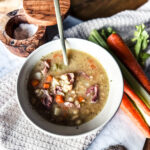 Bean soup placed in a creamy white bowl. There is a gold spoon placed into the bowl. IN the background are carrots and celery.