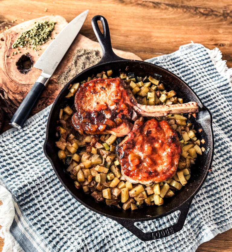 2 pork chops placed in a cast iron skillet on top of potatoes and onions. The backdrop is wooden with a blue towel