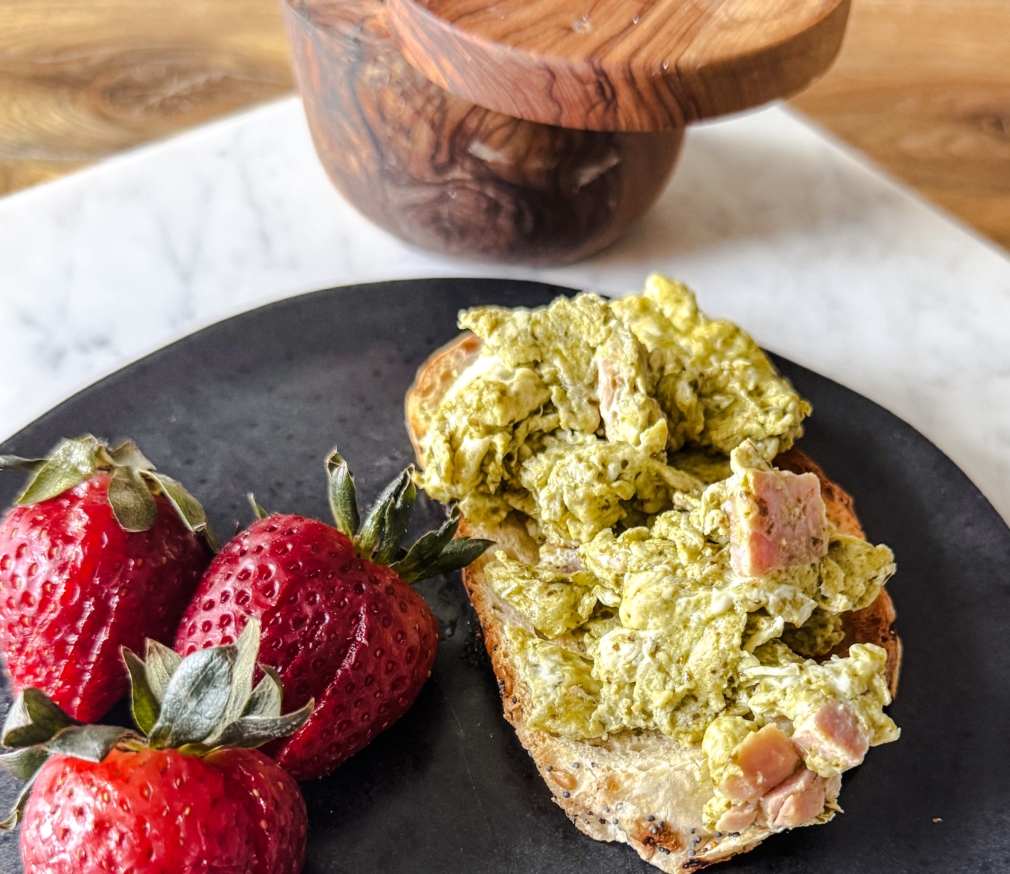 Pesto and ham eggs on top of a piece of toast with 3 strawberries sliced on the side. All placed on a black plate.