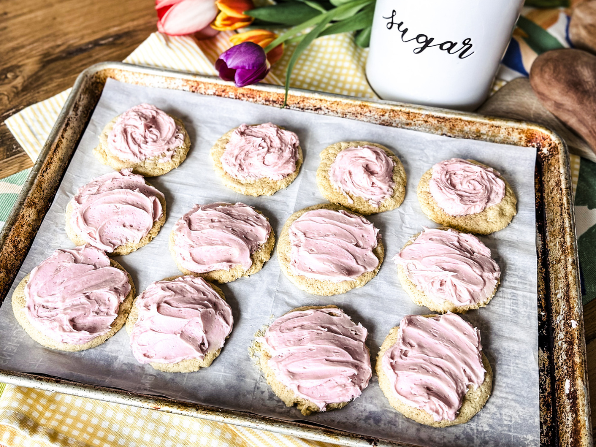 12 Lofthouse cookies that are placed on a parchment lined baking sheet. A wooden backdrop with tulips and a sugar jar.