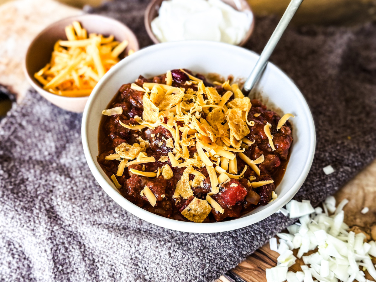 Chili placed in a white bowl with a wood and brown textured background. There is cheddar cheese and corn chips sprinkled on top. On the side is sour cream and cheese.