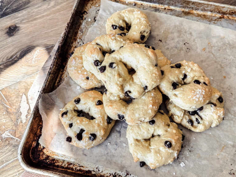 12 homemade chocolate chip sea salt bagels on a parchment lined baking sheet with a wooden background and orange tea towel behind.