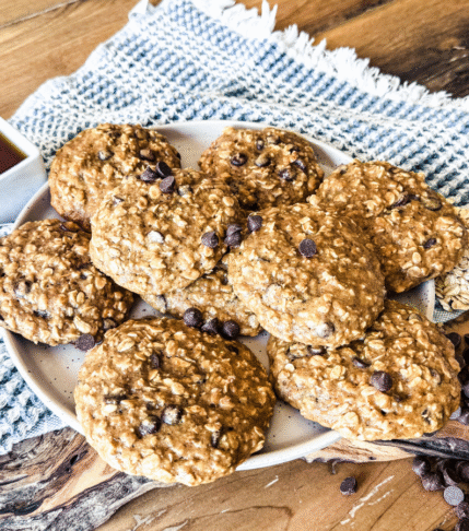 11 Breakfast cookies on an oval off white platter. Syrup in a small square dish to the left and chocolate chips and oats are on the right. The background is wood with a blue dish towel.
