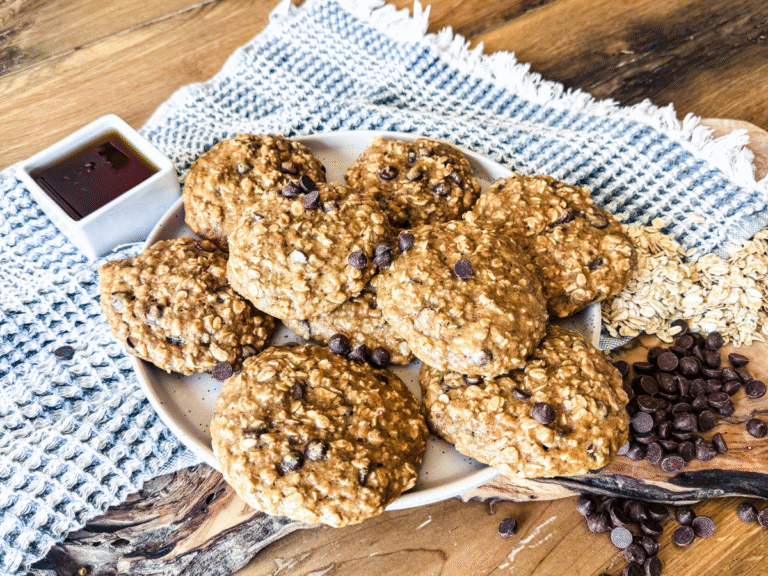 11 Breakfast cookies on an oval off white platter. Syrup in a small square dish to the left and chocolate chips and oats are on the right. The background is wood with a blue dish towel.