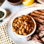 Homemade Baked beans in a small bowl on a plate with grilled pork chops to the right. A wooden back drop with tea towels under the plate and small bowls of seasonings in the background.