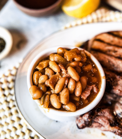Homemade Baked beans in a small bowl on a plate with grilled pork chops to the right. A wooden back drop with tea towels under the plate and small bowls of seasonings in the background.