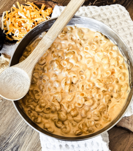 Macaroni and Cheese in a skillet with a wooden spoon laying across the top. The back drop is wooden with a white tea towel behind. Salt and shredded cheese are to the left.