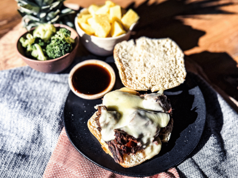 Hawaiian Beef Sandwich with Monterrey Jack Cheese on top and a bun top flipped open. A wooden background with orange tea towel. Pineapple chunks and broccoli to the top left.