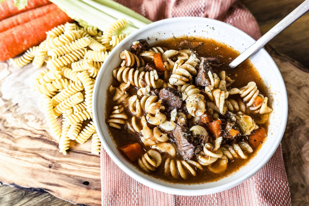 A white bowl of beef and pasta soup with a wooden backdrop and an orange tea towel behind. Pasta, carrots and celery to the left.