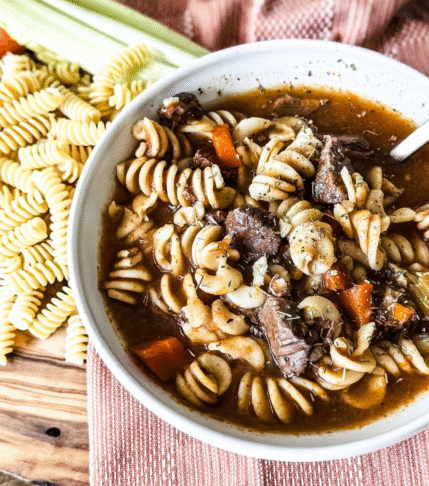 A white bowl of beef and pasta soup with a wooden backdrop and an orange tea towel behind. Pasta, carrots and celery to the left.