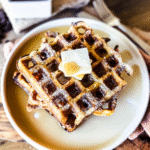 3 waffles piled high on a circular white plate. Butter in the middle and maple syrup poured over. A wooden backdrop with a rusty orange tea towel are in the background.