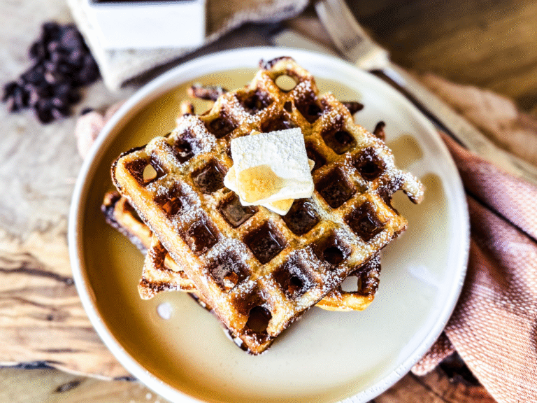 3 waffles piled high on a circular white plate. Butter in the middle and maple syrup poured over. A wooden backdrop with a rusty orange tea towel are in the background.