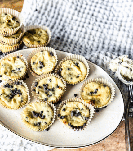 A wooden background with white textured tea towel. An oval off white platter rests with 9 mini cheesecake on top and a scoop with cheesecake batter to the right.