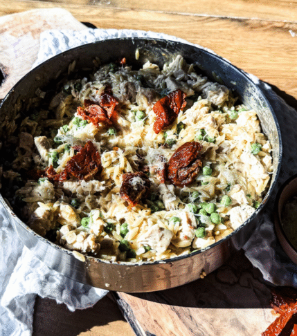 Skillet Sundried Tomato Chicken Orzo in a skillet with a gold handle. A wooden backdrop with a white textured towel behind.