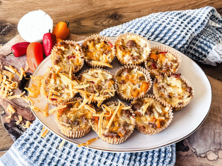 Spicy Egg Cups piled in an oval white platter with a blue dish towel and wood behind. Cheese is sprinkled over and peppers and onions rest to a size.