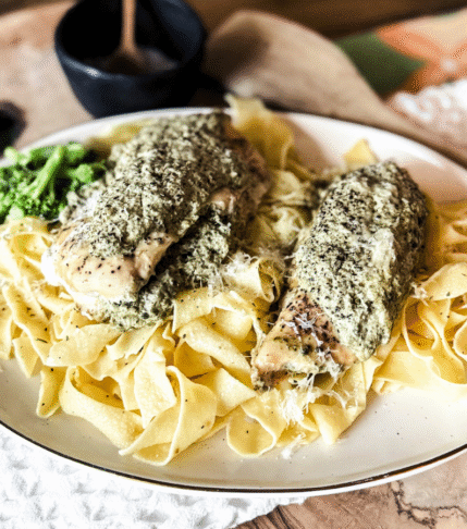 Wooden background with a white tea towel behind. Off white round plate with pappardelle pasta and stuffed artichoke chicken on top.