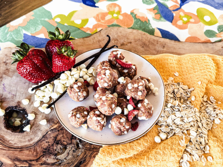14 strawberries and cream protein balls on a circular plate with strawberries on the top left with white chocolate chips and oats.