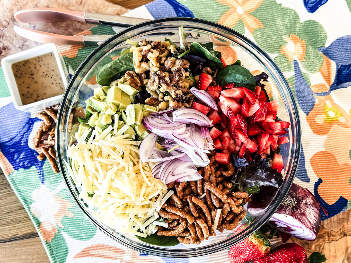 Strawberry Salad in a clear bowl. With a wooden and bright floral background and poppy seed dressing in a small square container to the left
