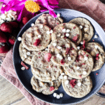 White Chocolate Strawberry Cookies on a black circle plate. A wooden background with an orange tea towel. Bright flowers and strawberries to the top left.