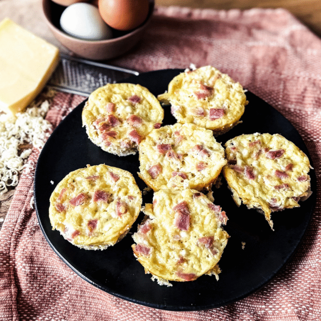 5 ham and gruyere egg bites on a black plate with an orange tea towel behind. A bowl of eggs sit to the top left with a brick of gruyere cheese and a grater.
