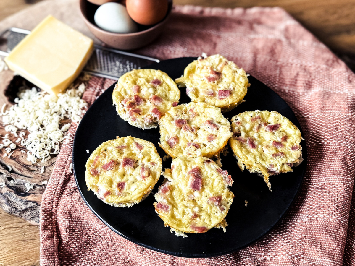 5 ham and gruyere egg bites on a black plate with an orange tea towel behind. A bowl of eggs sit to the top left with a brick of gruyere cheese and a grater.