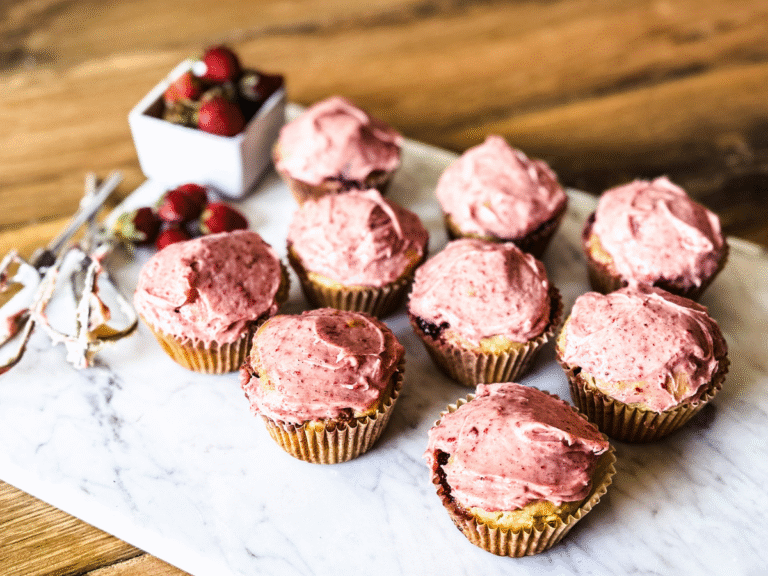 Strawberry Muffins on a marble platter with a small white cup to the top left. Batters with frosting are at the left.