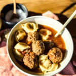 Sausage and Tortellini Soup in a white bowl with a gold spoon. A wooden backdrop with an orange tea towel are placed behind. Salt, onions and garlic are placed to the left.