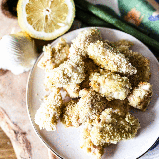 chicken bites piled on a white plate with lemon and garlic to the top left.