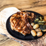 2 Pork Chop, potatoes and broccoli on a black round plate. A wooden background layered behind.