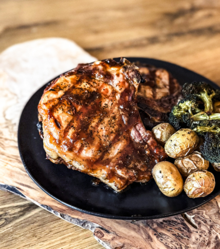 2 Pork Chop, potatoes and broccoli on a black round plate. A wooden background layered behind.